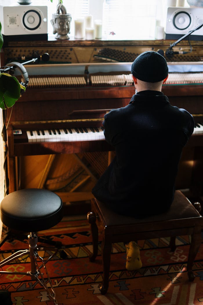 A man wearing a hat plays piano in a cozy home studio, surrounded by music equipment.